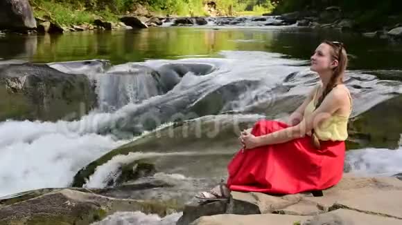 美丽的长发女孩穿着红色的裙子坐在山河瀑布背景上的岩石上夏日之夜安宁视频的预览图
