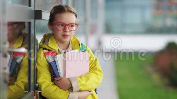 漂亮的女孩戴着眼镜和一件黄色夹克捧着书和微笑学校附近的女学生第一天上学视频的预览图