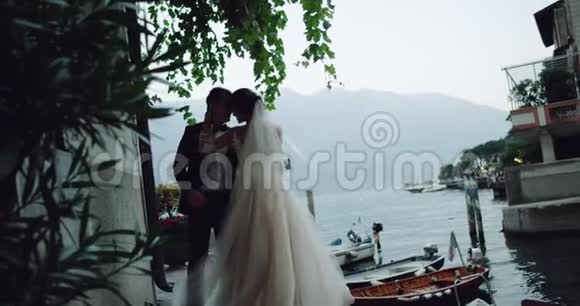 漂亮的新娘穿着长长的裙子新郎在迷人的地方亲吻和拥抱旁边有湖和船视频的预览图