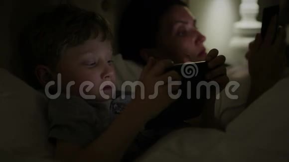 母亲和儿子躺在床上在黑暗中阅读他们的电子设备视频的预览图