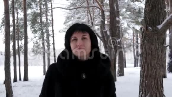 一个穿着毛皮外套的年轻黑发女人在霜冻的日子穿过一片雪林或公园看着镜头照相机视频的预览图