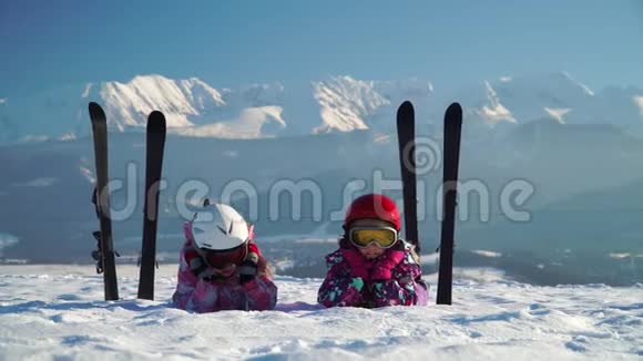 戴着头盔和护目镜的儿童躺在雪地上滑雪板靠近山脉背景视频的预览图