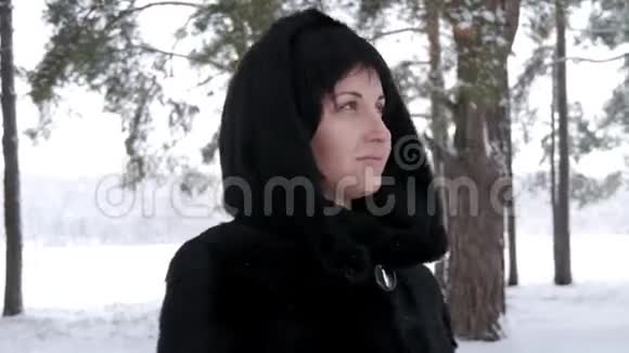 冬天下雪的时候一个穿着毛皮大衣的黑发女人站在森林或公园里看着远处摄像机移动视频的预览图
