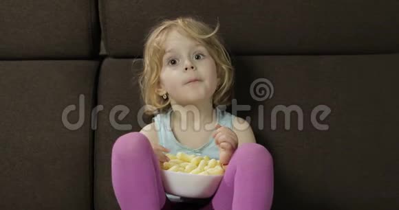 女孩坐在沙发上吃玉米泡芙儿童看电视品尝爆米花视频的预览图