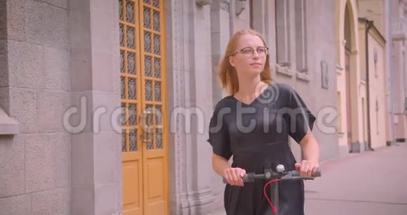 一位年轻漂亮的白种人女性戴着眼镜和连衣裙拿着踢踏板车看着她视频的预览图