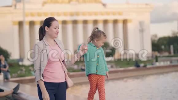 年轻的妈妈和贪玩的女儿在夏日公园的喷泉边散步视频的预览图