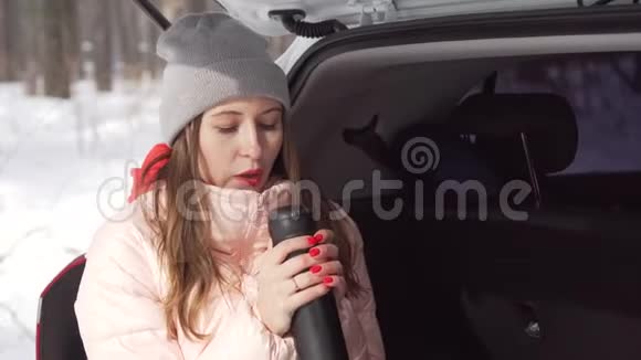 一个美丽的女孩坐在白色的车里在阳光明媚的冬天在森林里喝茶视频的预览图