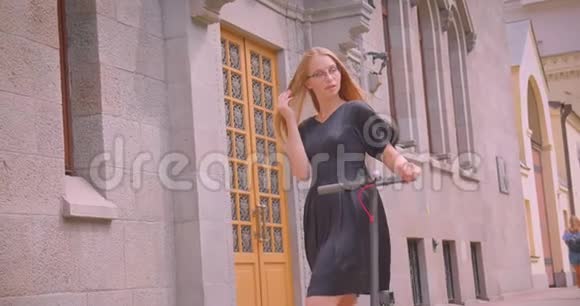 年轻漂亮的女性戴着眼镜穿着长头发拿着踢踏板车看着摄像机的特写照片视频的预览图