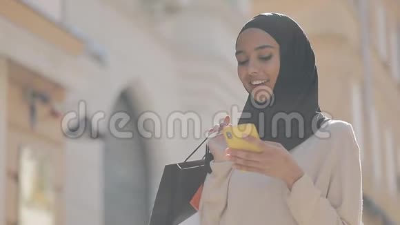 年轻的穆斯林妇女戴着头巾在寻找智能手机的销售手里拿着购物袋站在街上视频的预览图