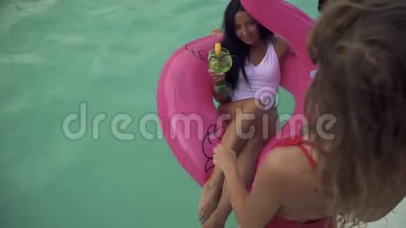 两个穿着泳衣的漂亮女朋友在游泳池里玩得很开心喝着鸡尾酒一个黑发女孩视频的预览图