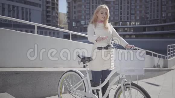 漂亮的金发女人穿着温暖的白色夹克站在城市的街道上骑着自行车看着镜头抚摸着她的头发视频的预览图
