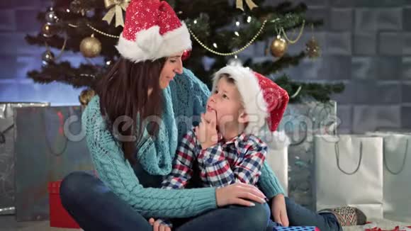 微笑的年轻妈妈和小可爱的孩子拥抱在一起坐在圣诞树旁聊天视频的预览图