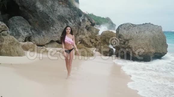 穿着泳装慢动作美丽苗条的女孩在海浪和大石头的沙滩上漫步视频的预览图