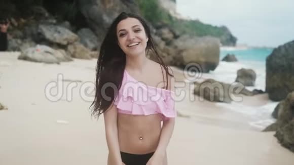 慢动作穿着泳装微笑的女孩在沙滩上散步背景是大石头和波浪Stealicam射击视频的预览图
