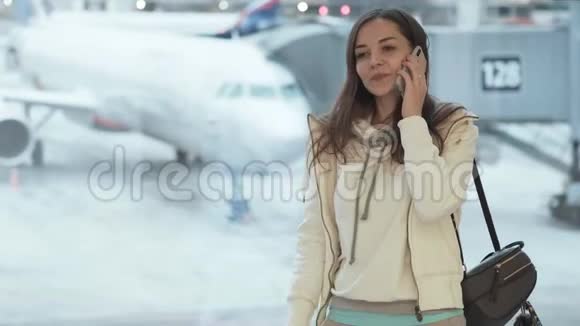 穿着运动服的漂亮女孩站在机场的窗口在飞机背景上打电话视频的预览图