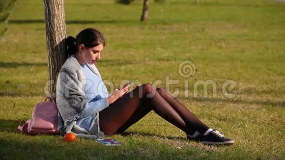穿着短裤和运动鞋的漂亮长腿漂亮的深褐色头发的女孩坐在树旁的绿草地上在电话里看着一些东西视频的预览图