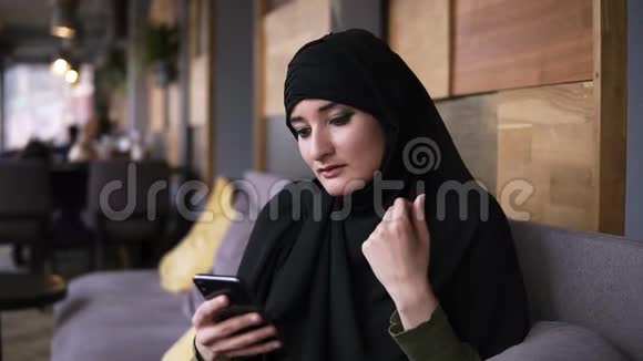 穆斯林妇女在咖啡馆使用她的智能手机聊天在线浏览社交媒体分享生活方式享受放松视频的预览图