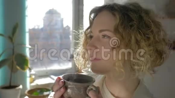 一幅梦中女孩在阳光明媚的窗户上用杯子特写喝热咖啡的肖像视频的预览图