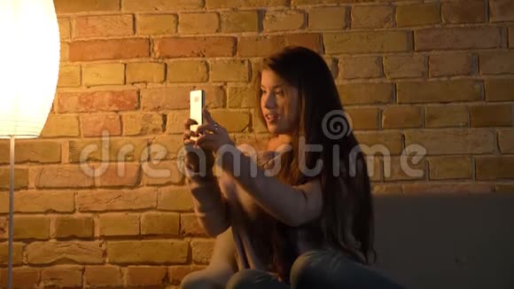 年轻的高加索女孩坐在沙发上在智能手机上拍照在舒适的家庭背景上微笑的肖像视频的预览图