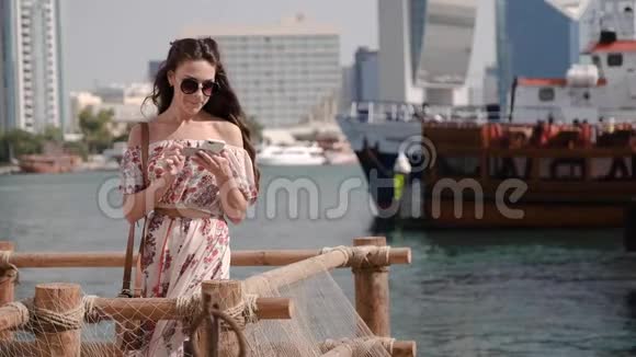 穿着连衣裙的微笑女孩在码头和船城发送短信视频的预览图