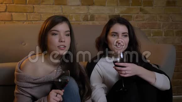 年轻的白种人女性朋友坐在地板上端着酒杯在舒适的家里看电影视频的预览图