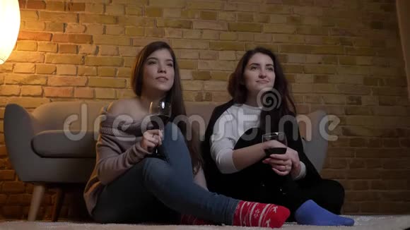 快乐的年轻白种人女性朋友坐在地板上端着酒杯在舒适的家里看电影视频的预览图