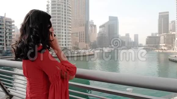 一个穿着鲜艳红色混血衣服的女孩在联合迪拜码头人工港旁边打电话视频的预览图