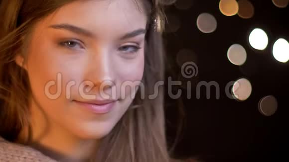 在模糊的灯光背景下年轻的白种人女孩在镜头中愉快地微笑的特写肖像视频的预览图