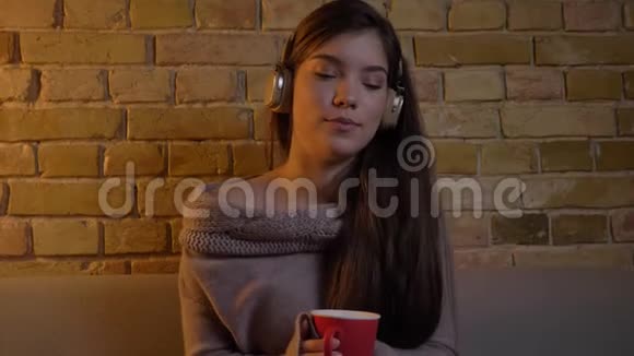 特写照片年轻的白种人女孩戴着耳机坐在沙发上红色的杯子放在布里肯墙背景上视频的预览图