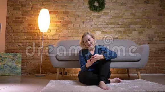 在一间舒适的公寓里一个年轻的白种人女人坐在地板上用手机拍摄她的特写照片视频的预览图