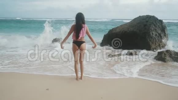 穿着泳装双臂张开的苗条女孩站在海滩上看着海浪稳重视频的预览图