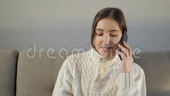 漂亮的女孩坐在家里的沙发上用她的电话说话视频的预览图