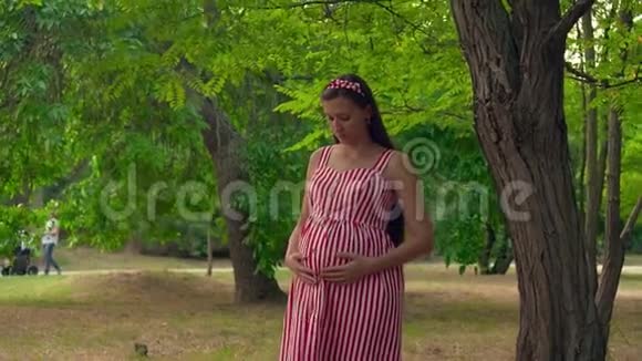 一个怀孕的女孩站在公园里一个穿着条纹白红色连衣裙的女孩站着把她的手放在肚子上视频的预览图