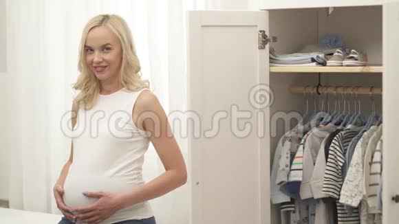 一个迷人的金发女郎站在打开的衣柜的左边穿着婴儿的衣服她把手放在肚子上视频的预览图
