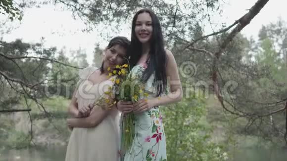 两个穿着夏装站在岩石地上开着野花看着森林的漂亮年轻女子的画像视频的预览图