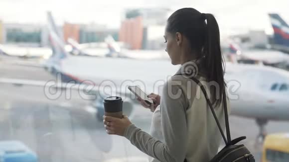 侧景女孩用手机和饮料外卖咖啡在机场背景的飞机在窗口视频的预览图
