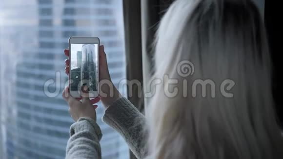 美丽的金发女人坐在床上用智能手机从窗户上拍了一张照片博客视频的预览图