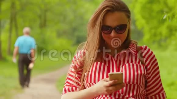 孕妇一只手握着电话快速键入信息对着镜头微笑漫步公园中弹视频的预览图