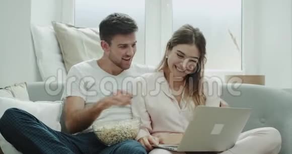 可爱的一对夫妇开心而微笑地吃着爆米花看着笔记本上的东西坐在沙发上舒适的风格视频的预览图