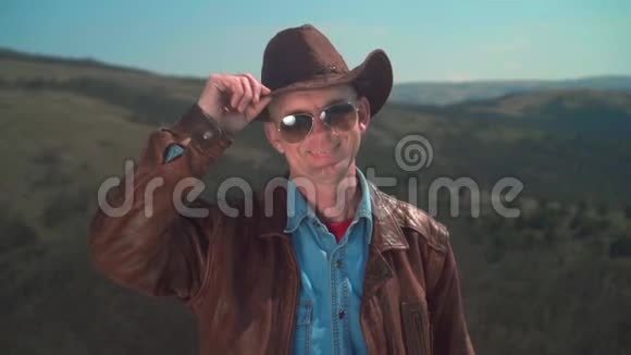 在山上一个男人戴着牛仔帽皮夹克眼镜一个人用手摸他的帽子视频的预览图