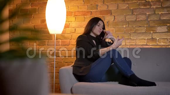 年轻的黑发白种人女孩坐在沙发上在家里的气氛中愉快地用手机交谈视频的预览图