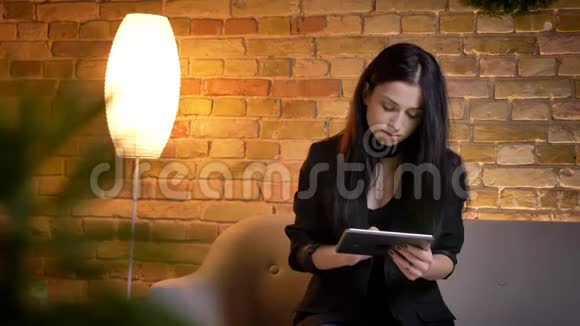 年轻的黑发白种人女孩坐在沙发上在家庭氛围中专心地与平板电脑合作的肖像视频的预览图