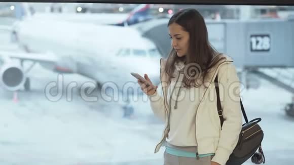 穿着运动服和背包的女孩站在机场的窗口在飞机背景上打电话视频的预览图