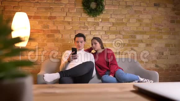 年轻的白种人夫妇坐在沙发上看着智能手机在家庭氛围中尽情地评论着内容视频的预览图
