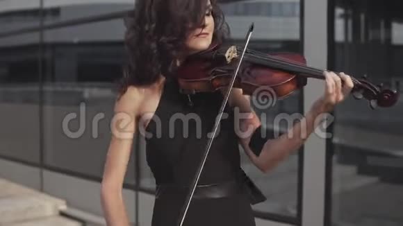 靠近优雅的女人穿着黑色连衣裙在玻璃大厦附近拉小提琴视频的预览图