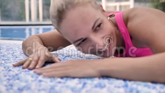 穿着粉红色泳衣的金发女郎笑着躺在游泳池的蓝色边视频的预览图