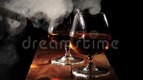豪华白兰地在黑色背景的棕色木桌上放两个带有金色干邑的玻璃杯抽雪茄的人抽烟视频的预览图