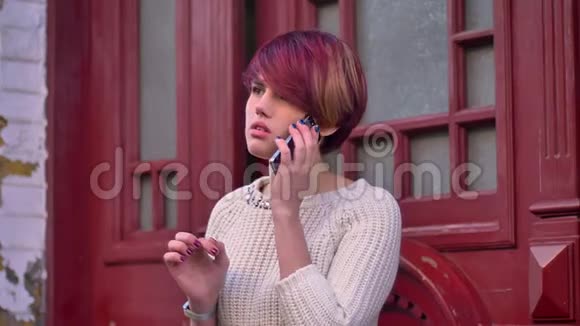 年轻的白种人粉红色头发的女孩在红色的门背景上用心地用手机说话的肖像视频的预览图