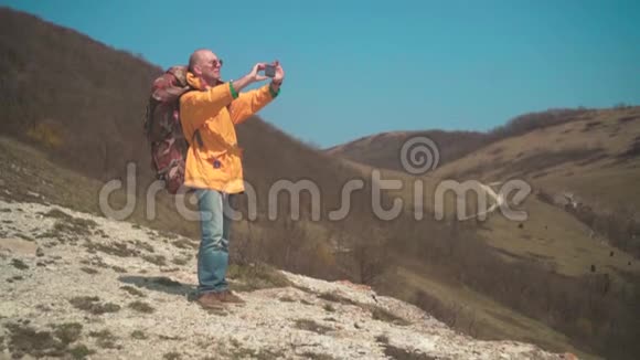 一个穿着黄色夹克和眼镜的人站在山上欣赏风景在电话里拍照视频的预览图