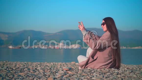 一个年轻的女孩戴着眼镜在春天的海滨穿着外套一个女孩在电话里拍了张照片视频的预览图